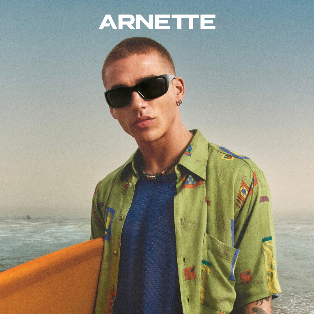 Das Bild zeigt einen jungen Mann mit Sonnenbrille und Surfbrett vor dem Meer.