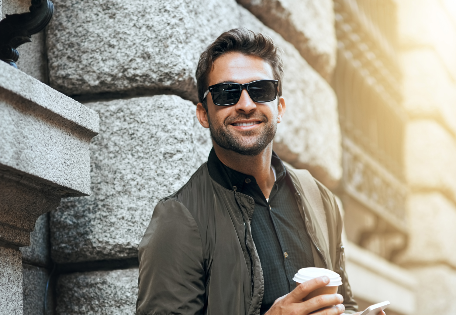 Das Bild zeigt einen Mann mit Sonnenbrille, der an einer Hauswand lehnt. Er hat einen Kaffee in der Hand.