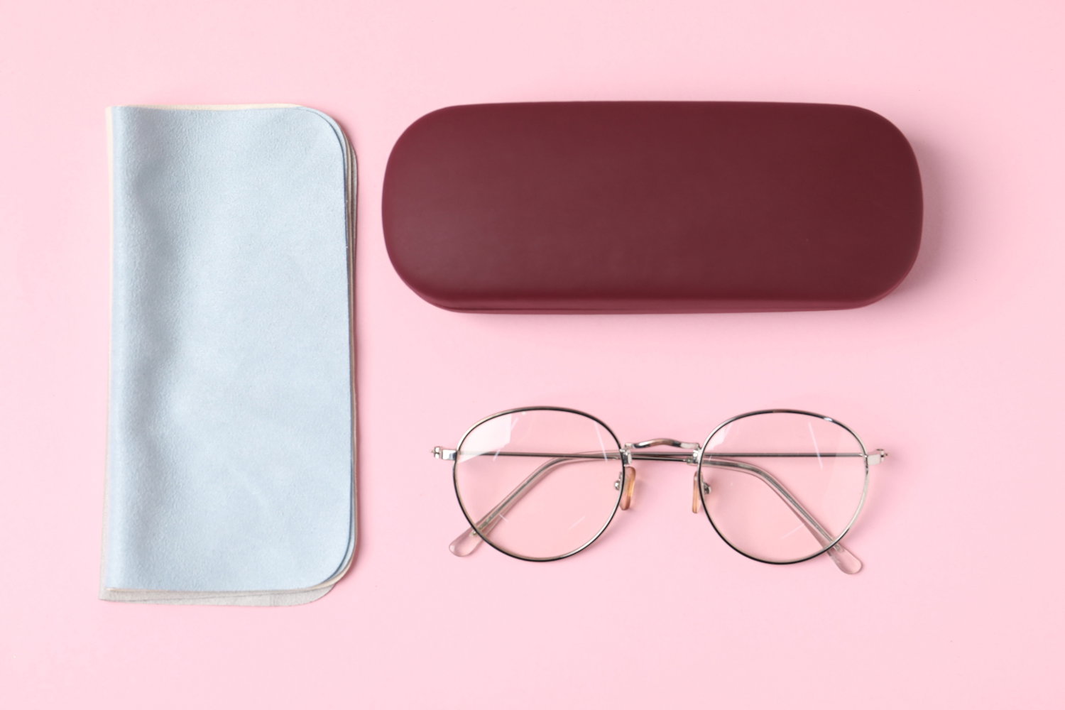 Das Bild zeigt eine Brille, ein Etui und ein Mikrofasertuch vor einem rosa Hintergrund.